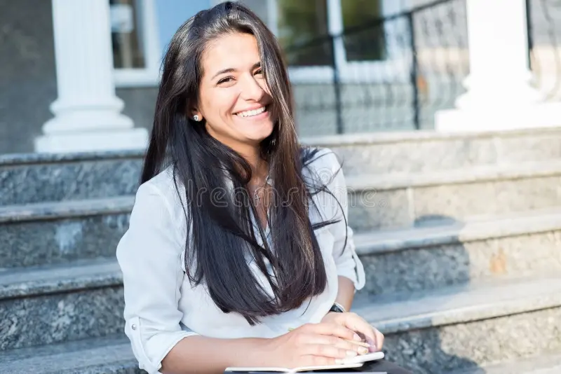 Home portrait beautiful young indian girl business woman smiling sitting steps office building holding notepad white 158880029 (1)